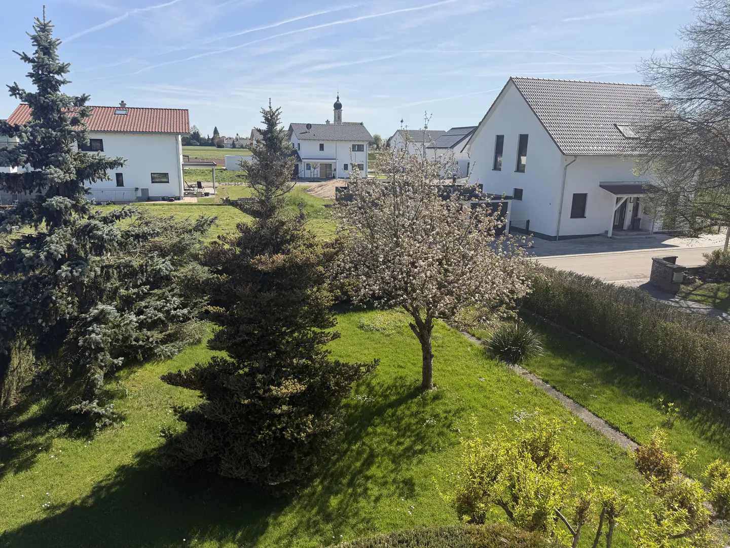 A sunny view of a green lawn with trees, white houses, and a church steeple in the background.
