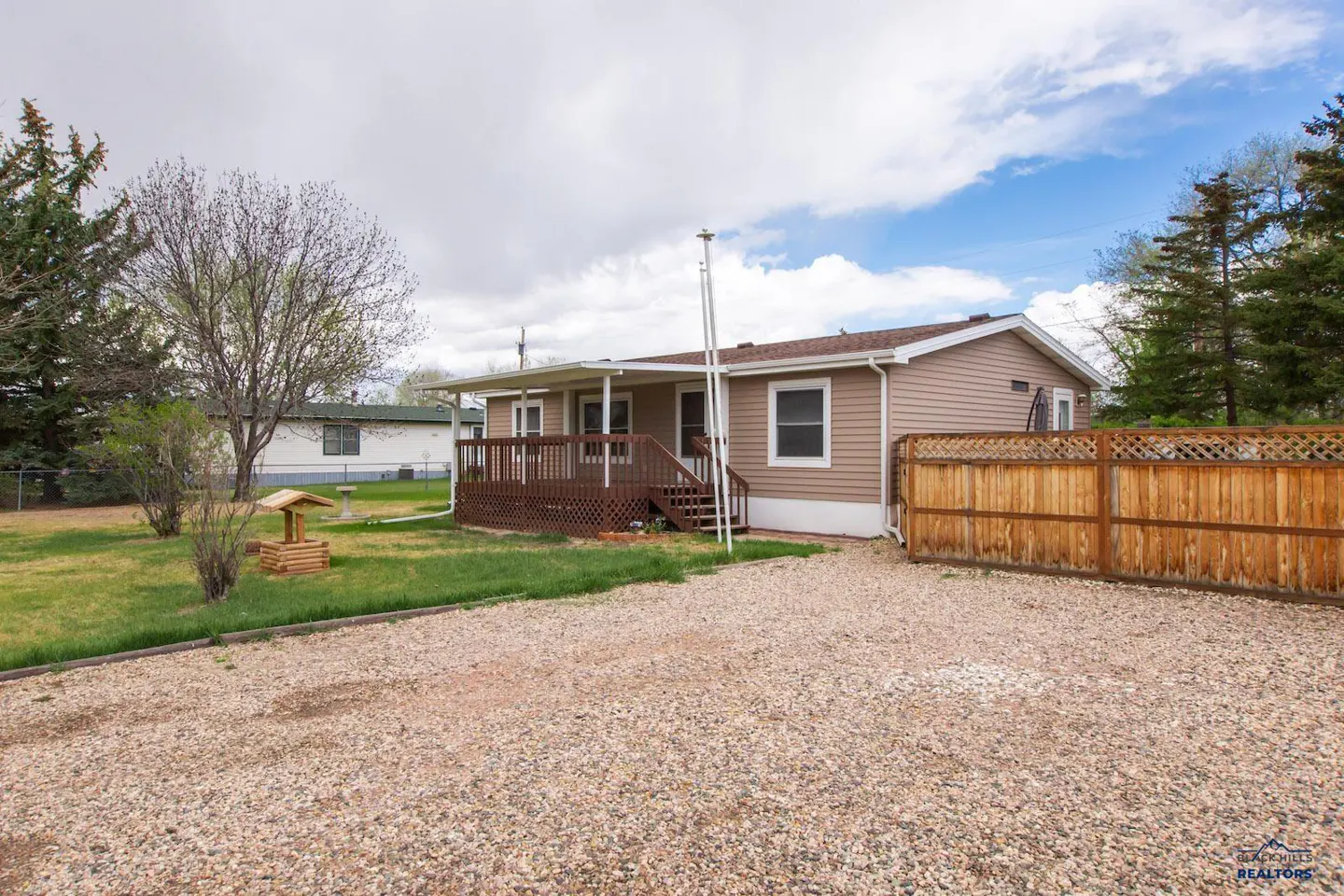 Tan single-story home with a brown deck and wooden fence, surrounded by a gravel driveway and green lawn.