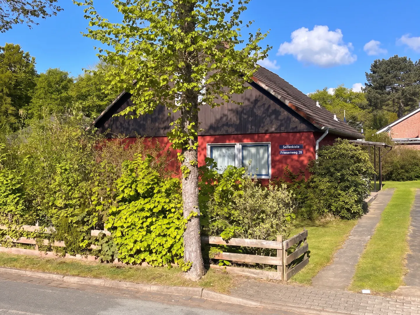 A red house with a dark roof, surrounded by green trees and bushes, under a blue sky with white clouds.