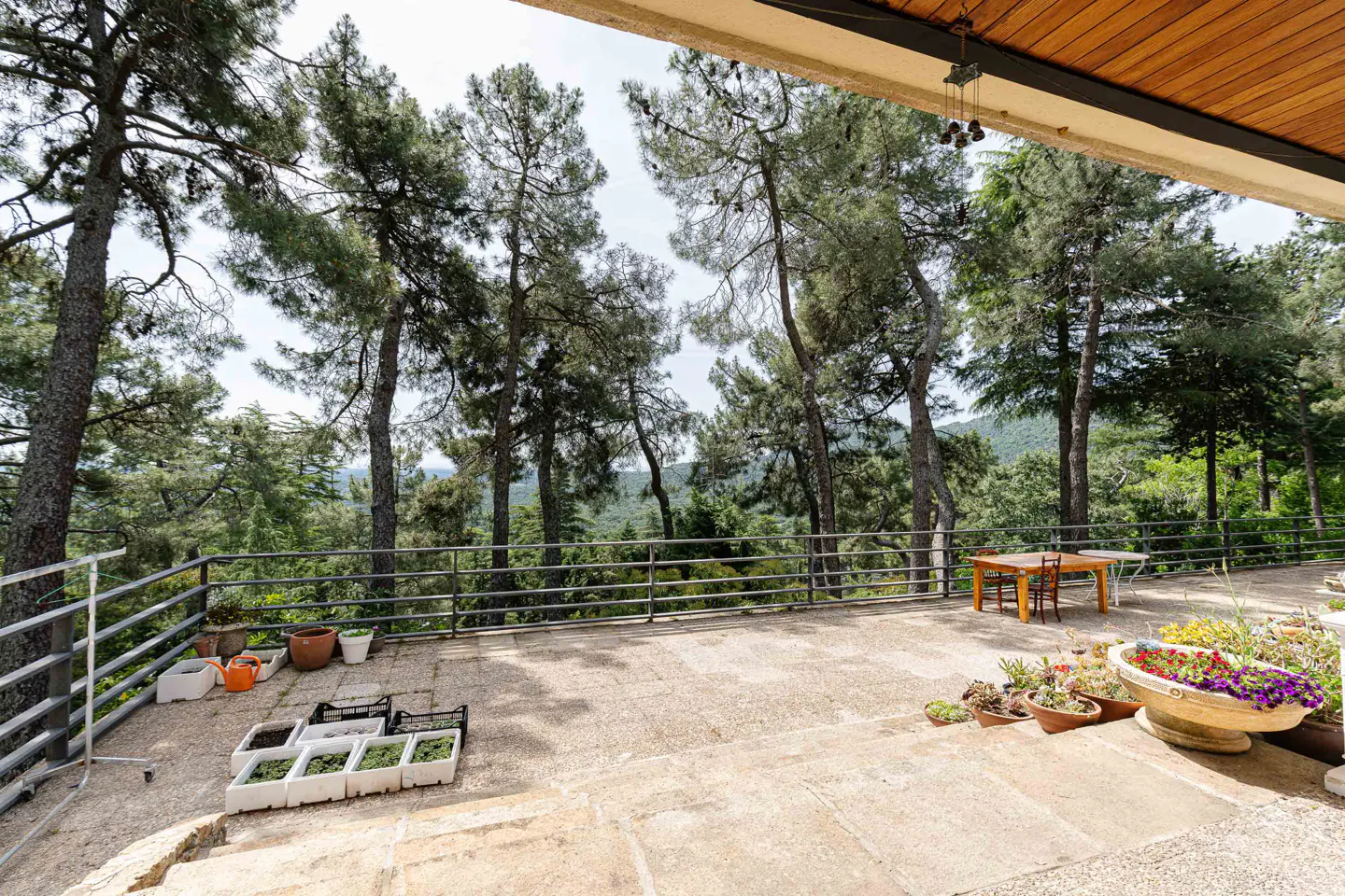 Stone patio with metal railing overlooks a forest. Potted plants and a wooden table with chairs are visible.