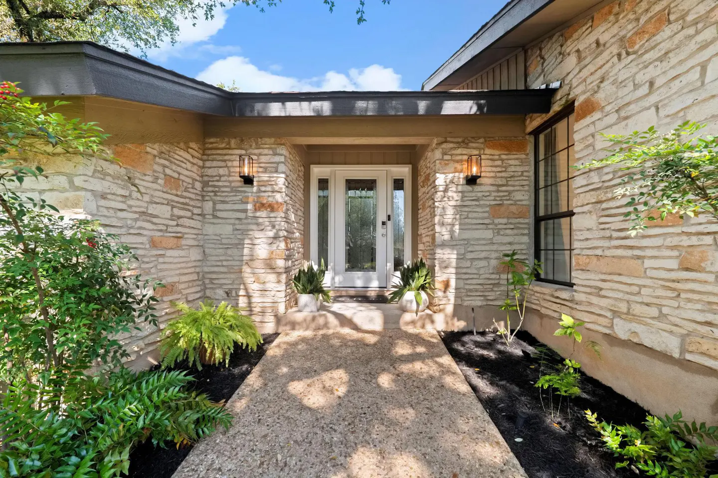 Exterior of a stone house with a white front door and a concrete walkway. Green plants are on either side of the walkway.