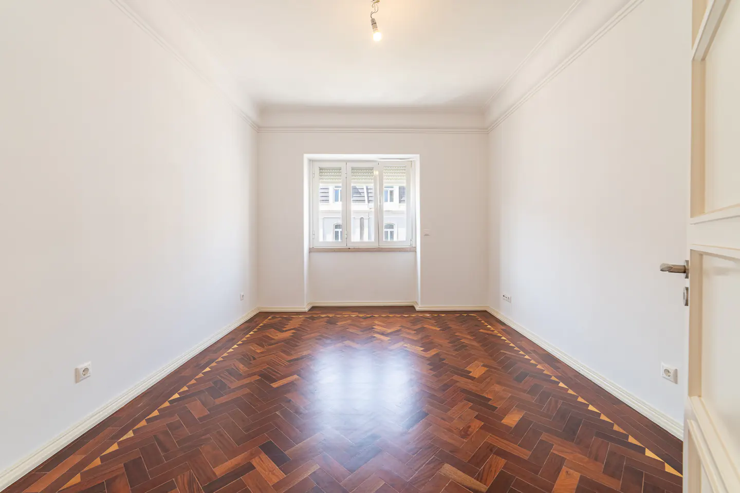 Empty room with white walls, herringbone wood floor, and a window with white shutters. A door is slightly open on the right.