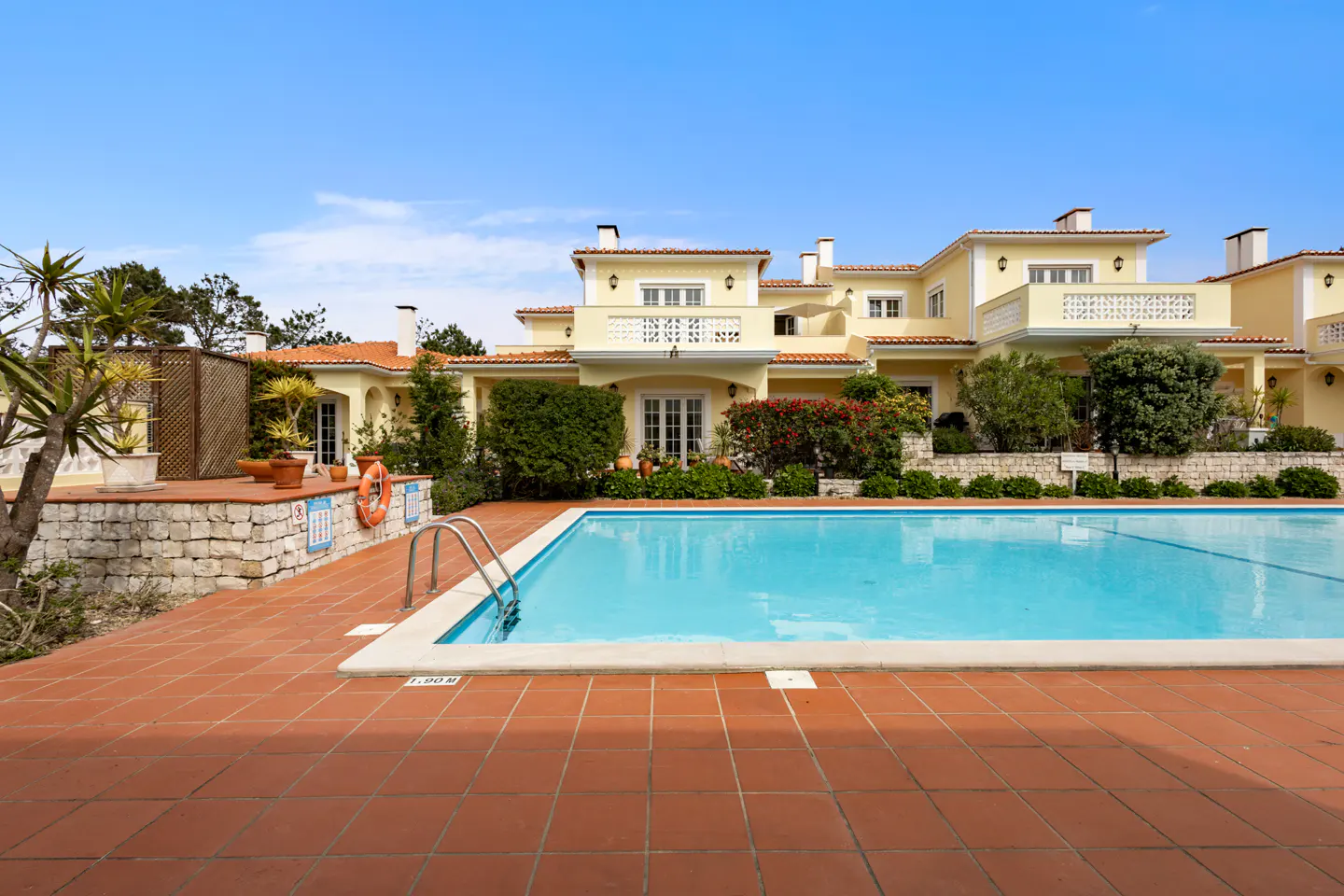 A yellow villa with a red tile roof overlooks a bright blue swimming pool with a red tile deck.
