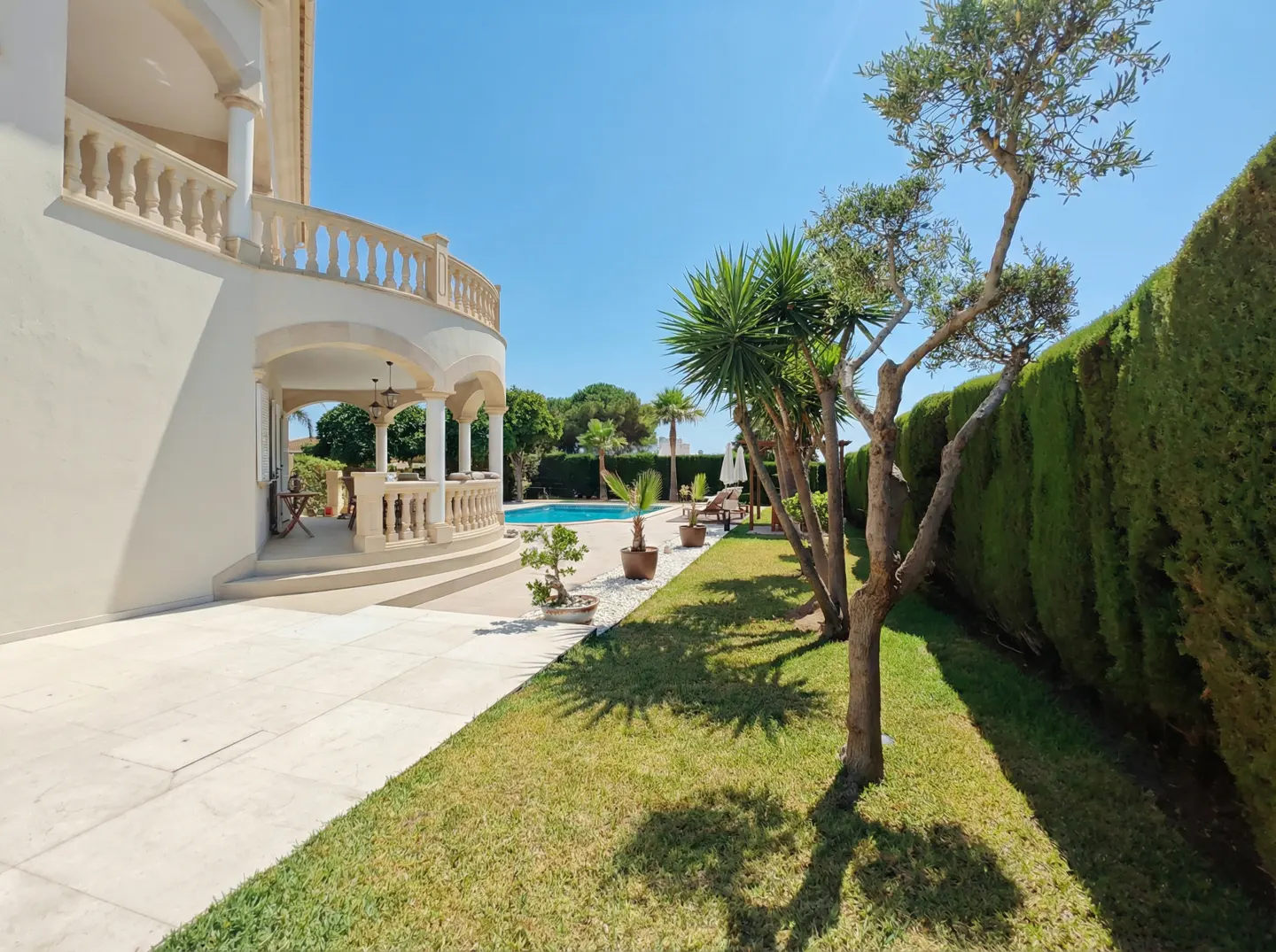 Exterior view of a luxury home with a pool, manicured lawn, and trees under a clear blue sky.