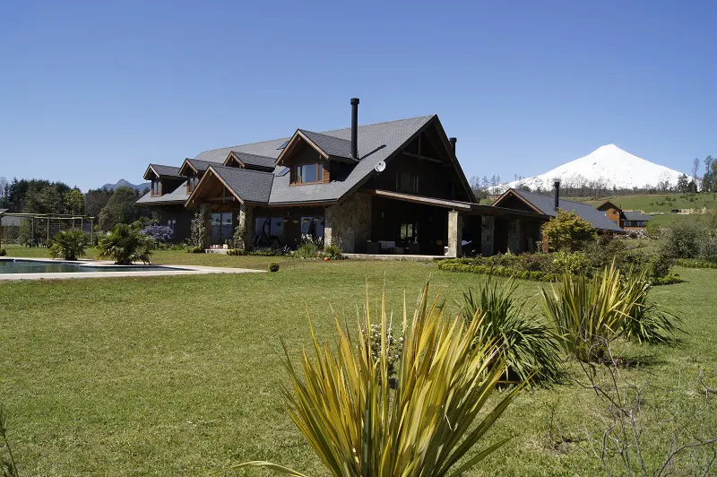 A large, brown, stone house with a gray roof sits on a green lawn under a blue sky, with a snow-capped mountain in the background.