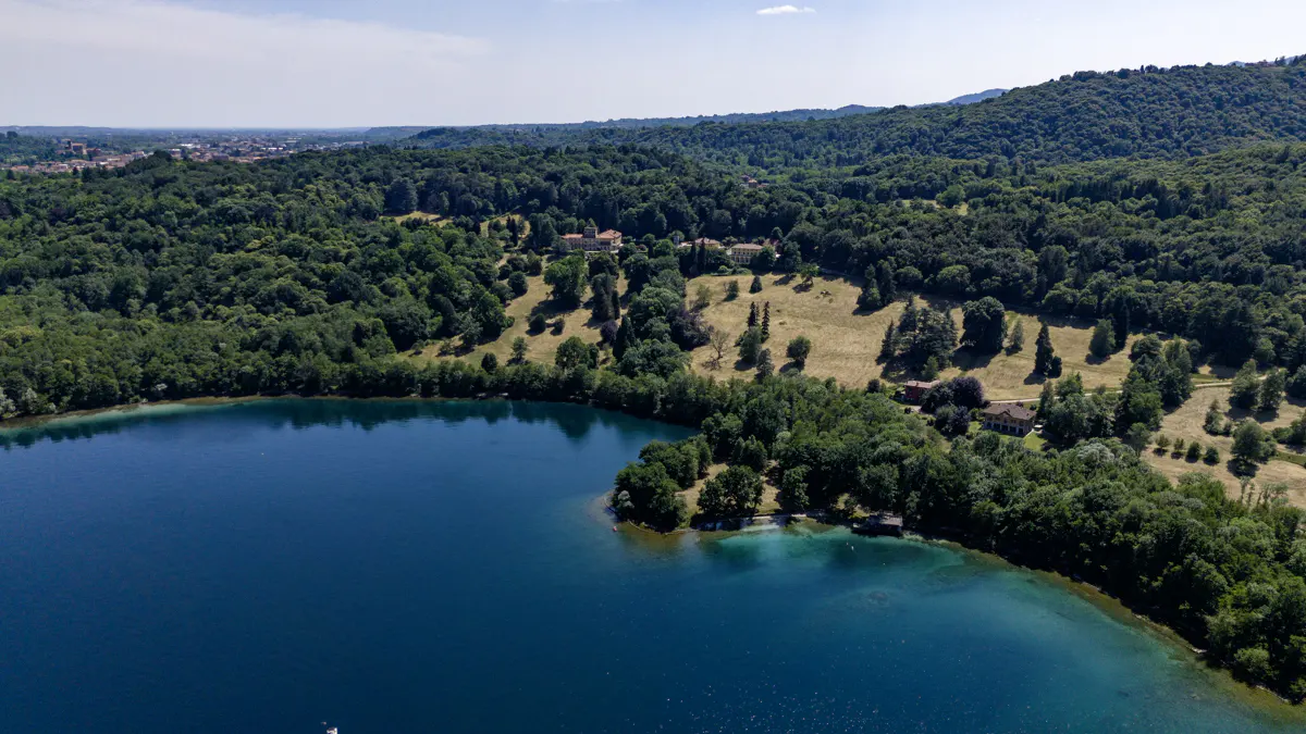 Aerial view of a deep blue lake surrounded by lush green forests, rolling hills, and secluded villas under a clear sky.