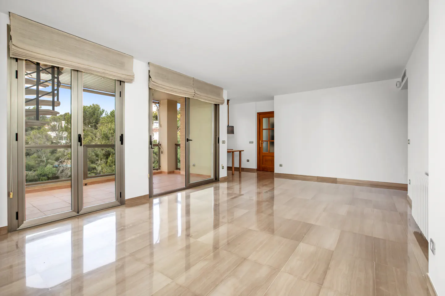 Bright, empty room with beige tile floor, white walls, and two sliding glass doors to balconies. A wooden door is on the right.