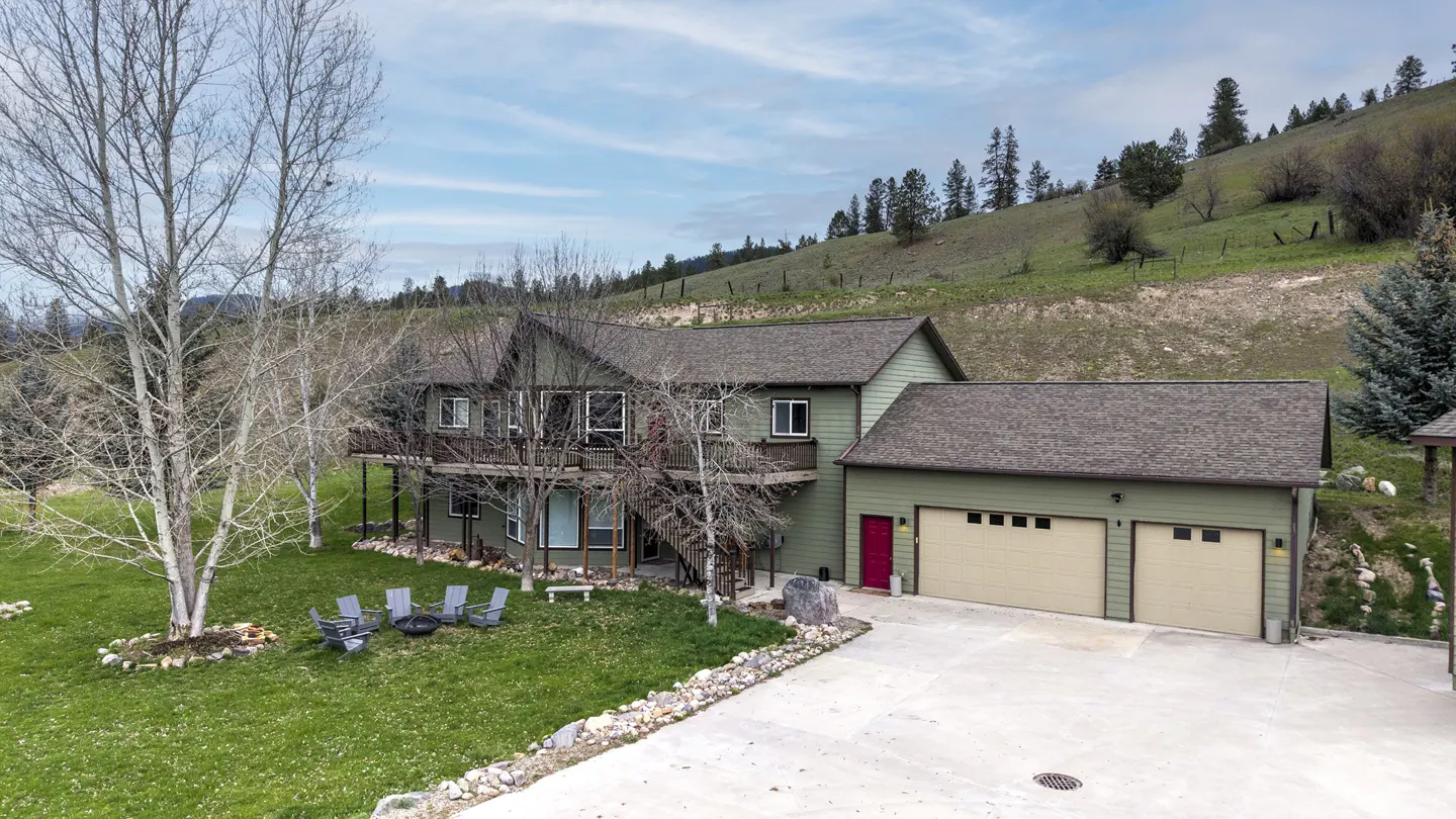 Two-story green house with a two-car garage, red door, and a green lawn with a fire pit and chairs.