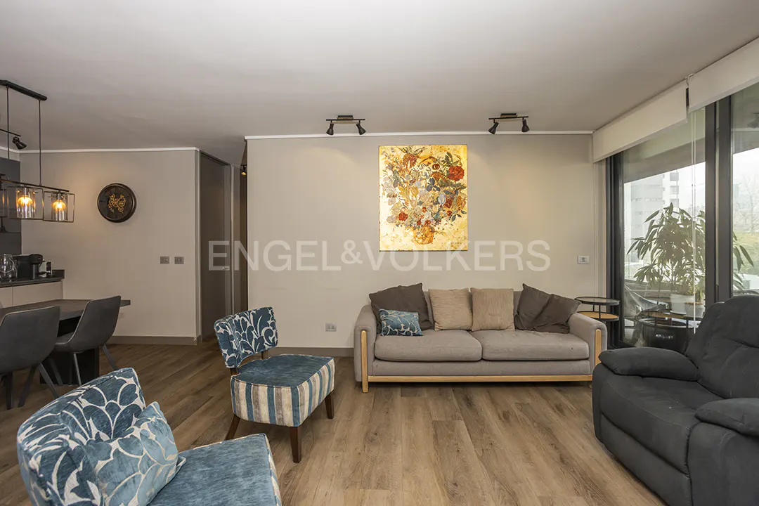 Living room with wood floors, gray sofa, blue chairs, and floral art. Large windows offer natural light.