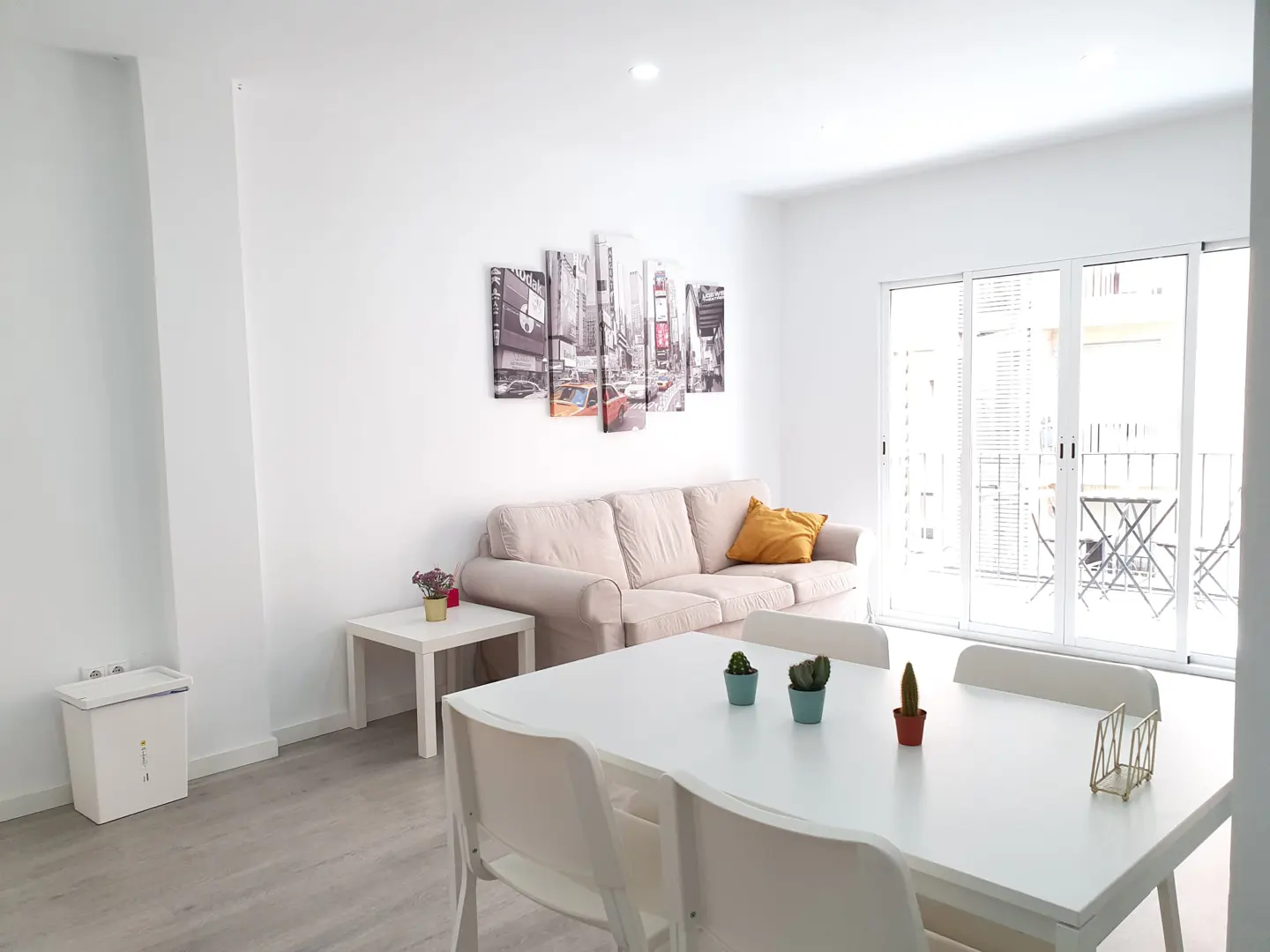 Bright living room with a white dining table, beige sofa, city-themed wall art, and a balcony with outdoor seating.