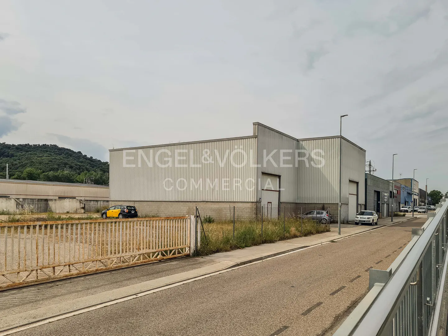 Exterior view of a light-colored industrial warehouse building with a metal fence and parked cars on a paved street.