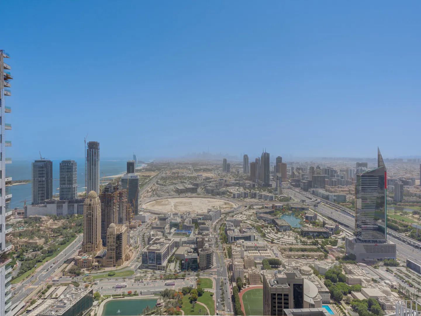 High-angle view of a sprawling city skyline with modern skyscrapers, a large circular construction site, and a clear blue sky.