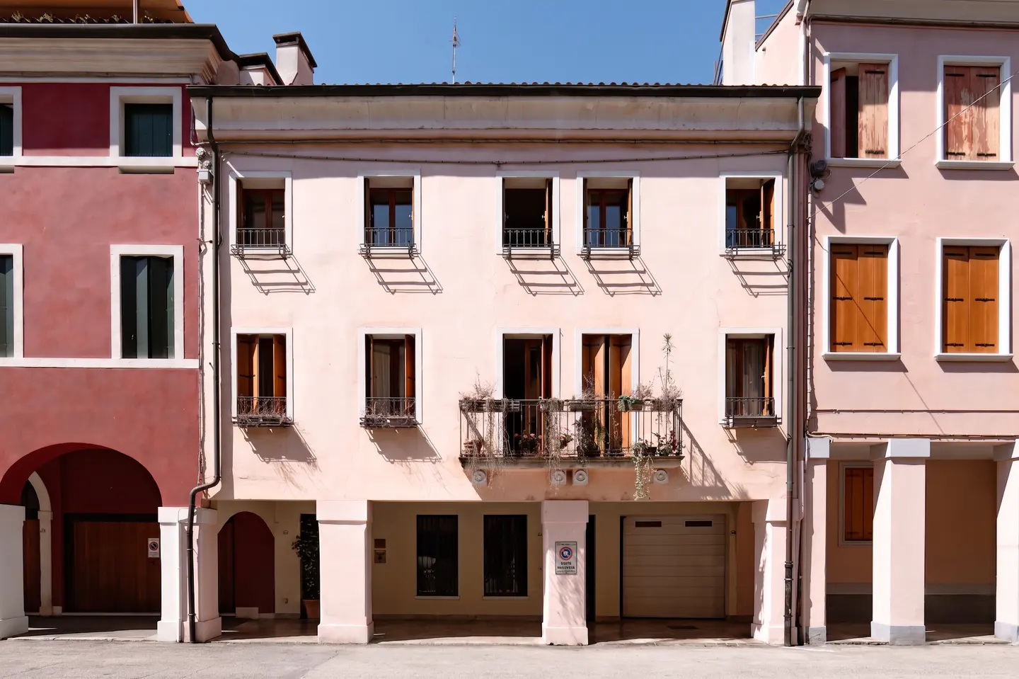 Facade of a three-story building with pink walls, arched entrances, and windows with dark shutters. A balcony with plants is on the second floor.
