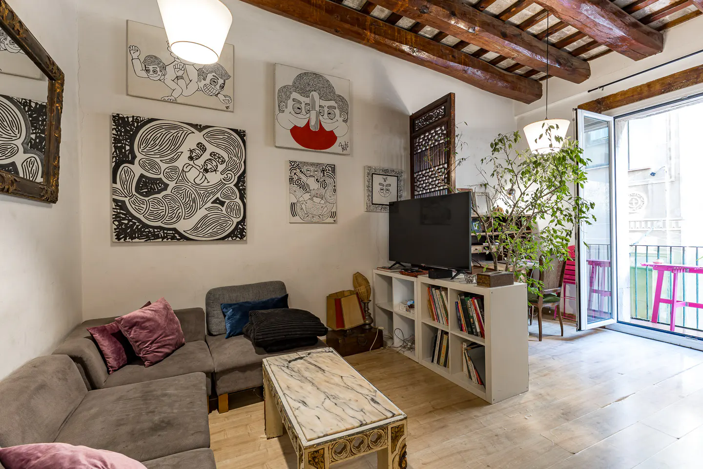 Living room with gray sectional sofa, marble coffee table, and white shelves with TV. Balcony with pink table and chairs visible. Artwork on walls.