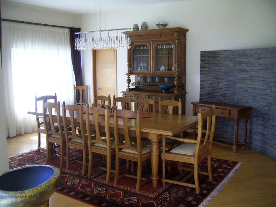 A dining room with a long wooden table, chairs, and a large wooden hutch. A crystal chandelier hangs above the table. A red patterned rug covers the floor.