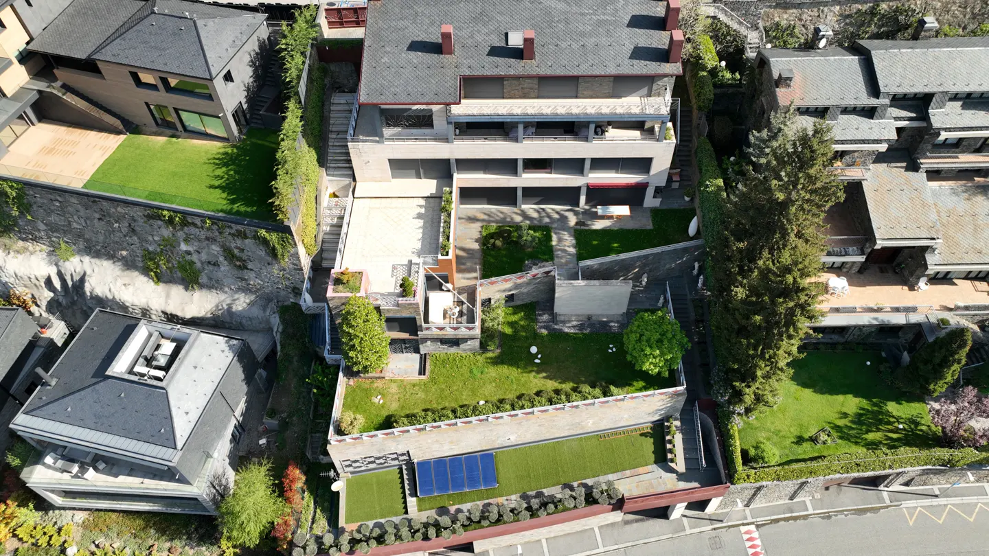 Aerial view of a multi-story modern house with terraced gardens, stone walls, and solar panels on a sloped residential lot.