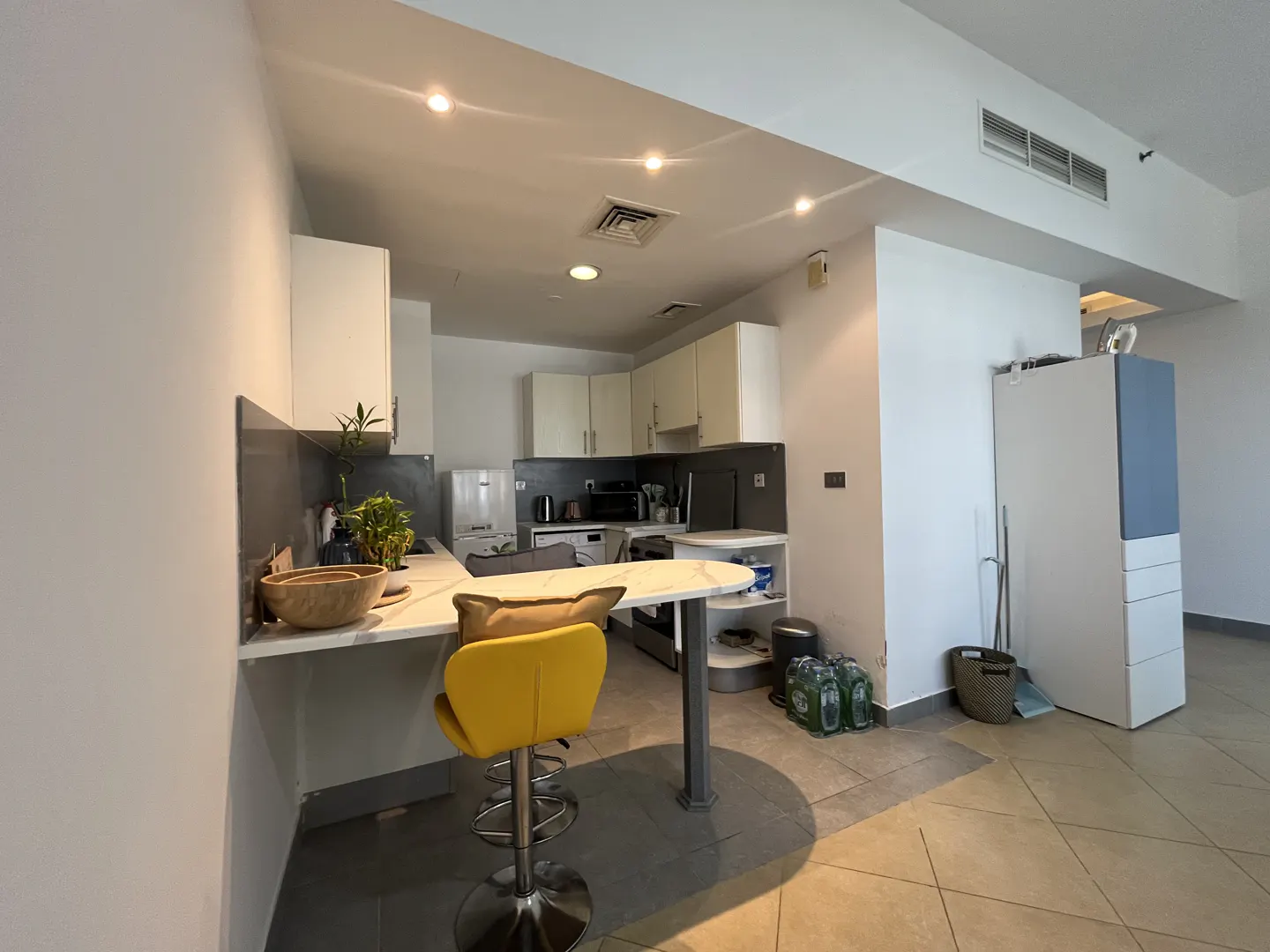 A bright kitchen with white cabinets, a marble countertop, and a yellow bar stool. Appliances are visible in the background.