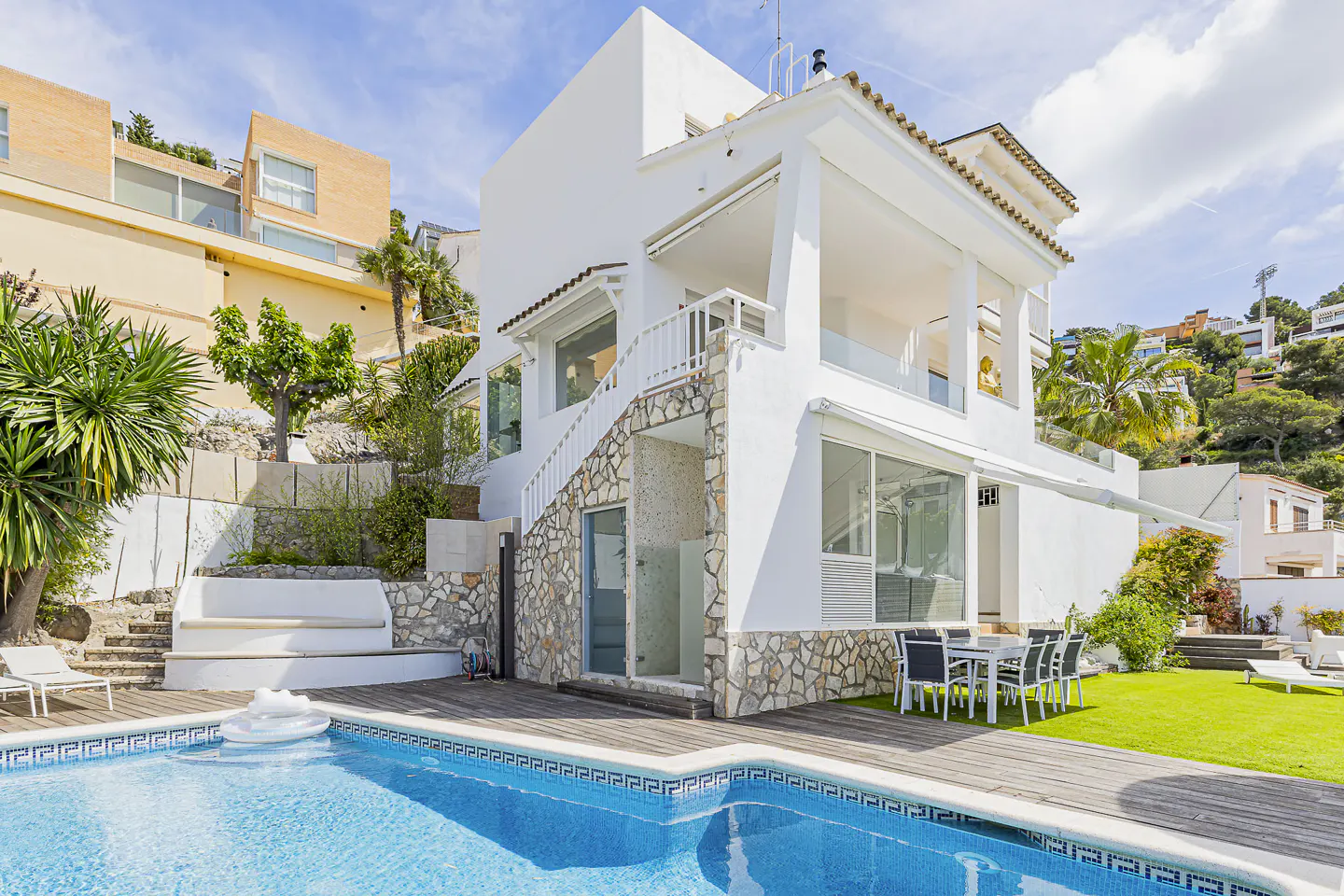 A modern white house with a pool and patio on a sunny day. The pool is blue and the patio has a table and chairs.