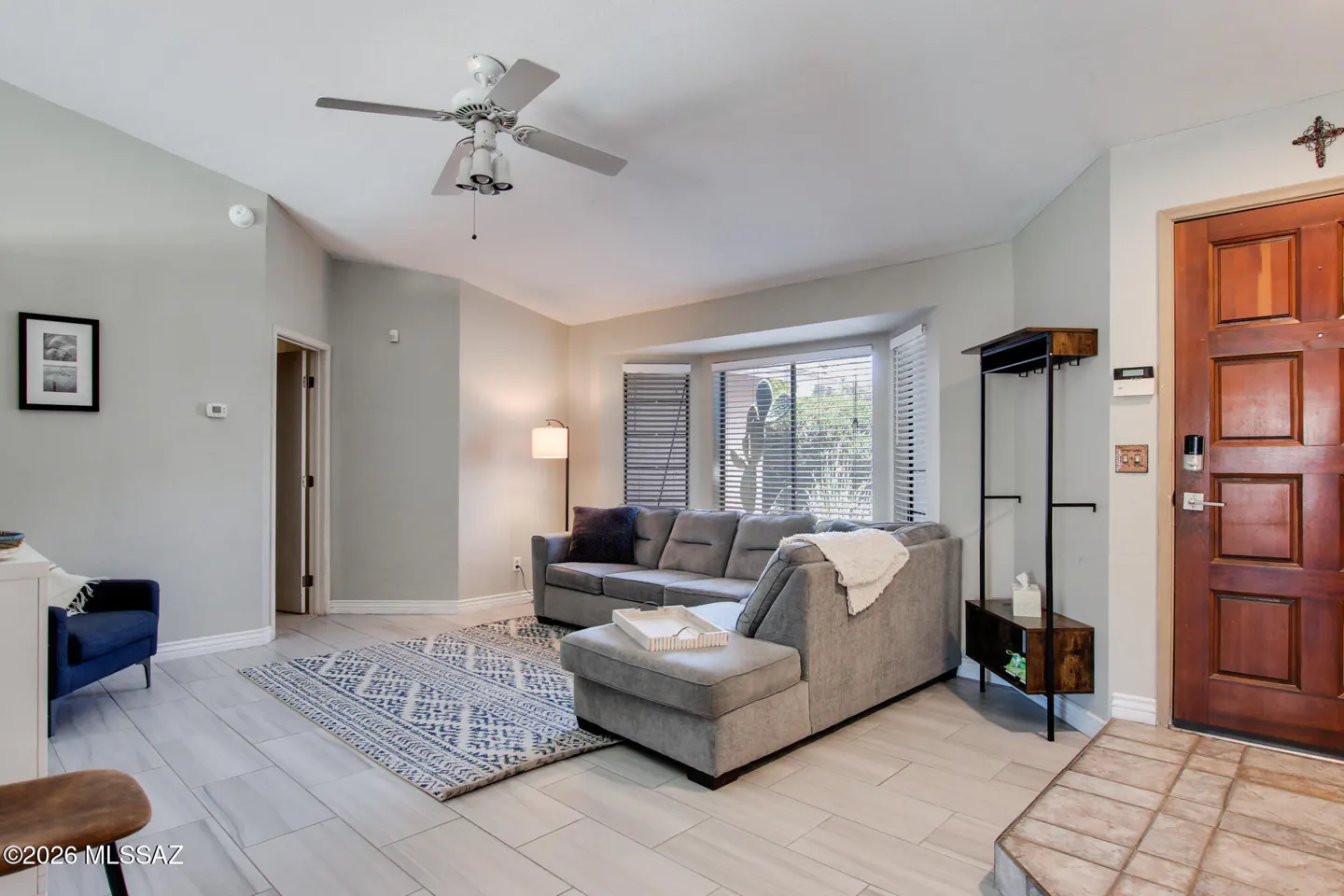 Living room with gray sectional sofa, patterned rug, and wood door. A ceiling fan hangs above.