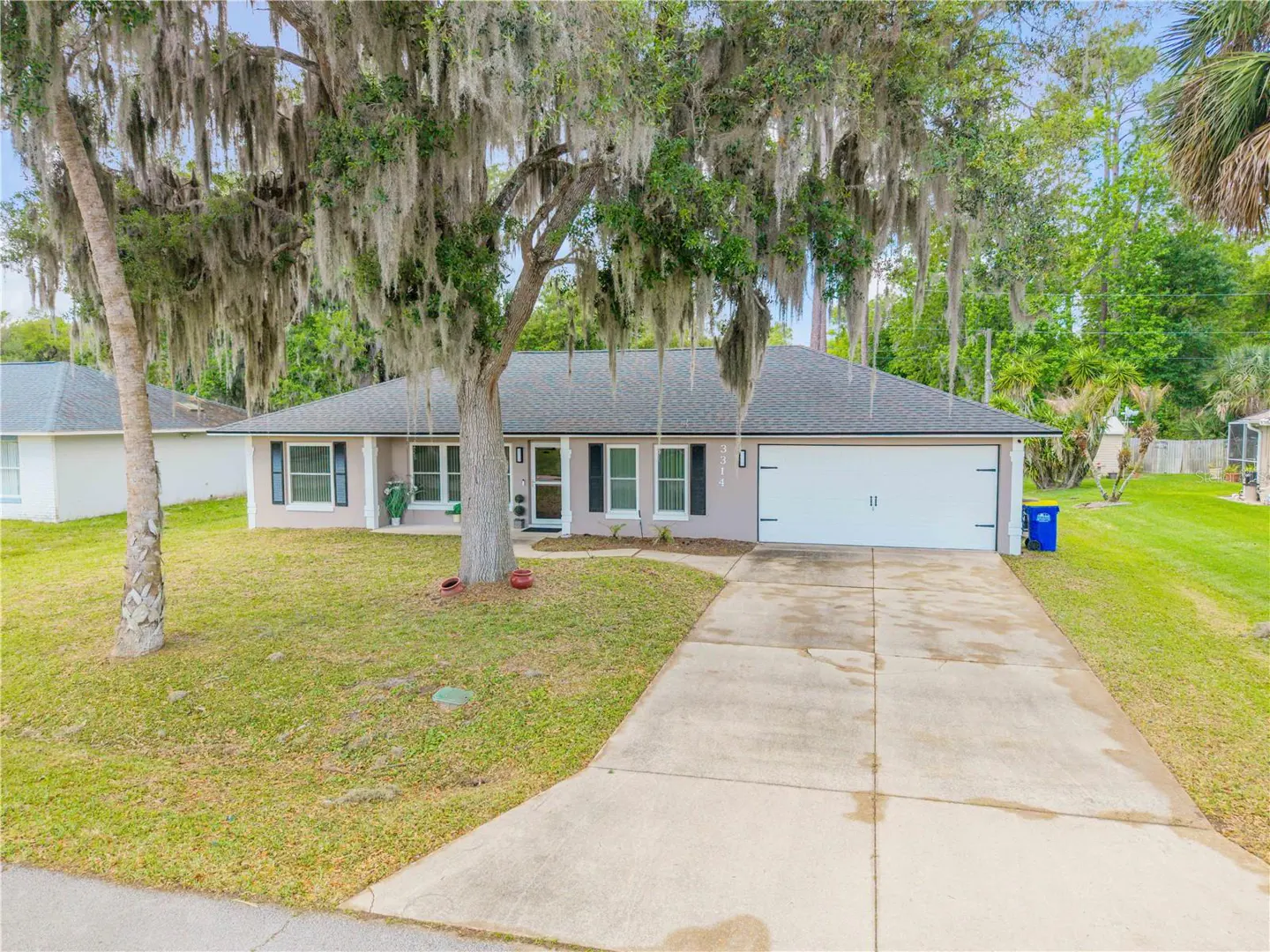 A single-story house with a gray roof, light pink walls, and a white garage door. A large tree with Spanish moss hangs over the front yard.
