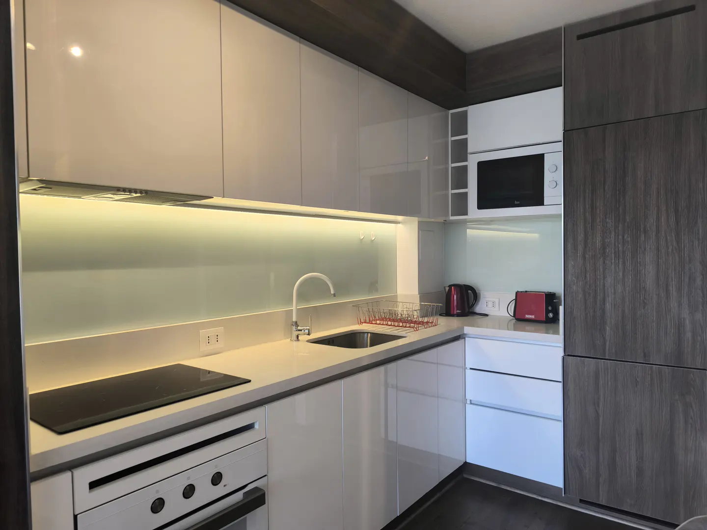 Modern kitchen with white cabinets, stainless steel sink, black cooktop, and gray wood-grain refrigerator.