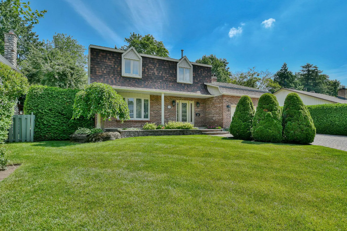 A brick house with a brown roof, white trim, and a green lawn under a blue sky.