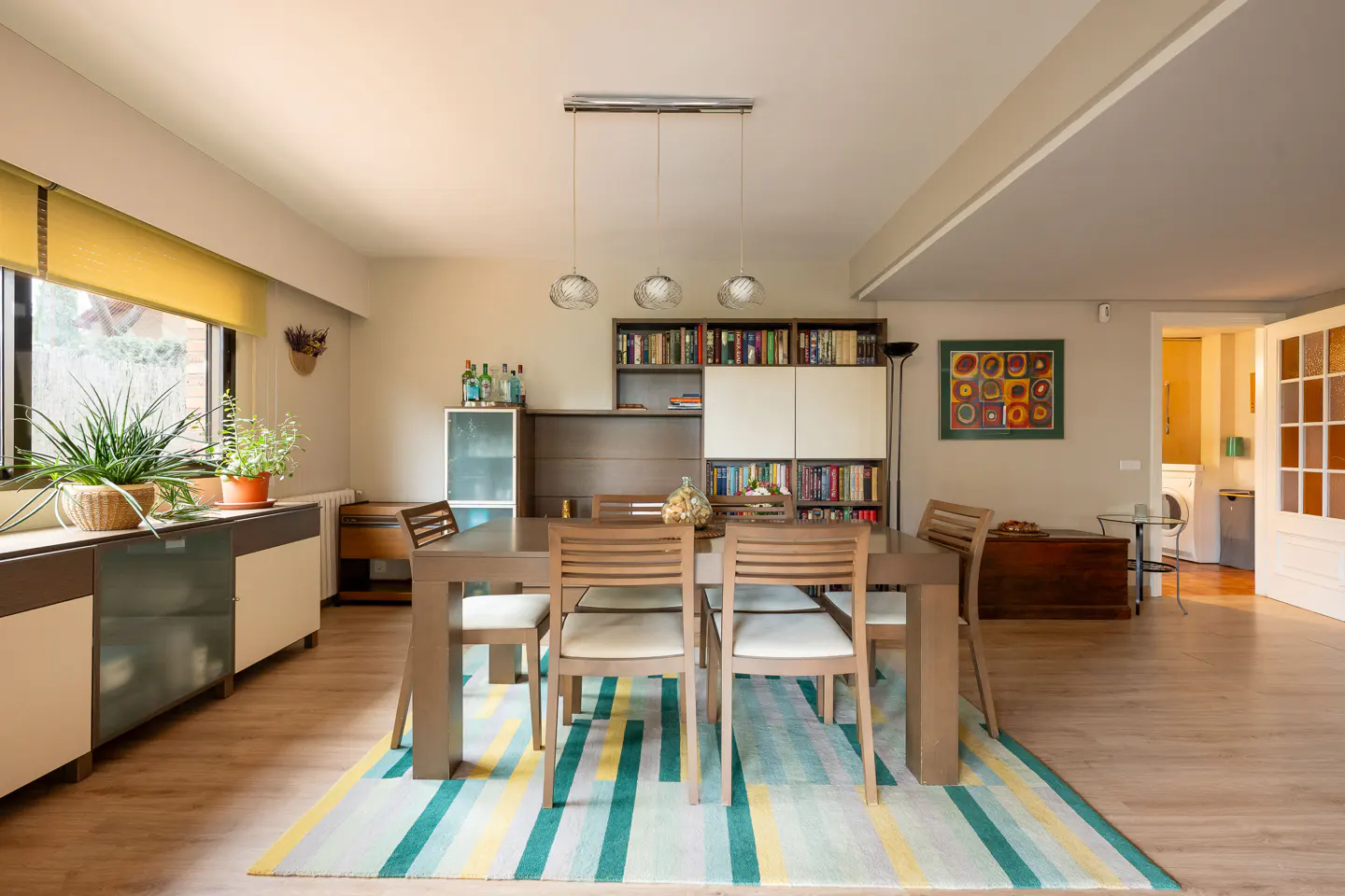 Bright dining room with a wooden table, chairs, and a colorful striped rug. Bookshelves and plants add a homey touch.