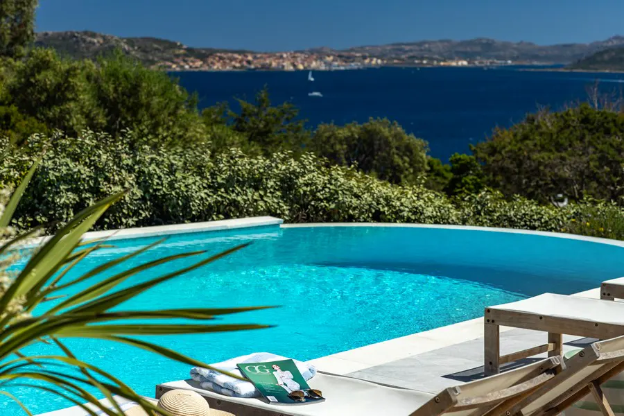 Outdoor pool with blue water, lounge chairs, magazine, and ocean view with distant buildings under a clear blue sky.