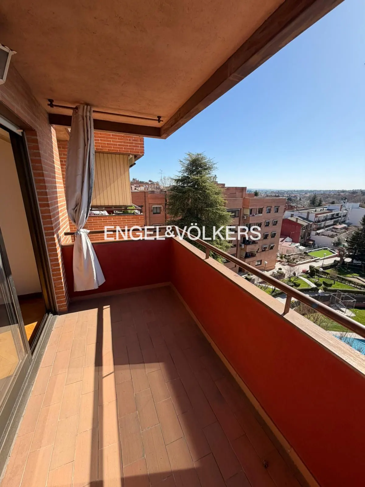 A balcony with terracotta tiles and a red railing overlooks a cityscape under a clear blue sky. A gray curtain is tied to the brick wall.