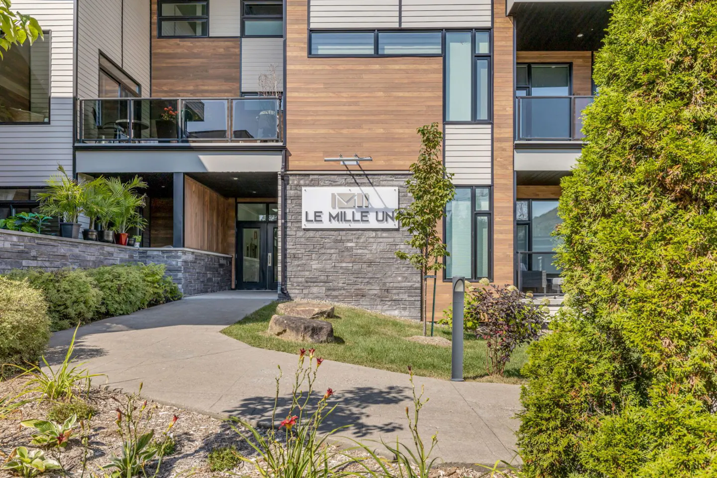 Exterior view of "Le Mille Un" apartments with a stone and wood facade, a walkway, and green landscaping.