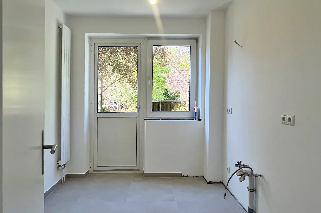 Bright, empty room with white walls, gray tile floor, and a white door/window combo looking out to trees. A radiator is on the left wall.