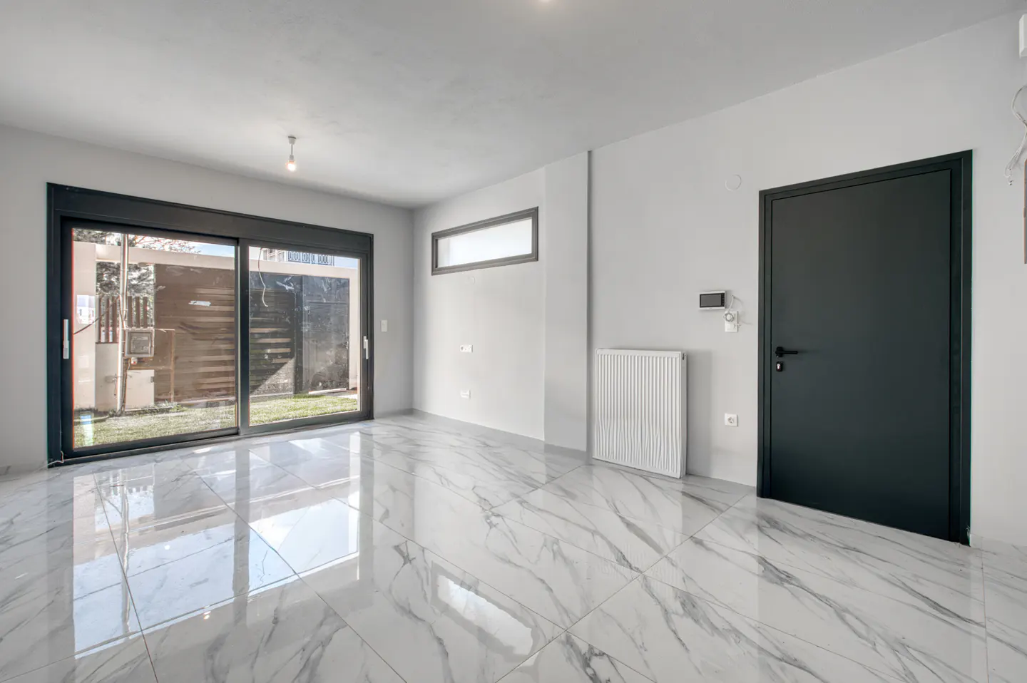 Bright, empty room with white walls, marble floors, and a black door. Sliding glass doors lead to a fenced yard.