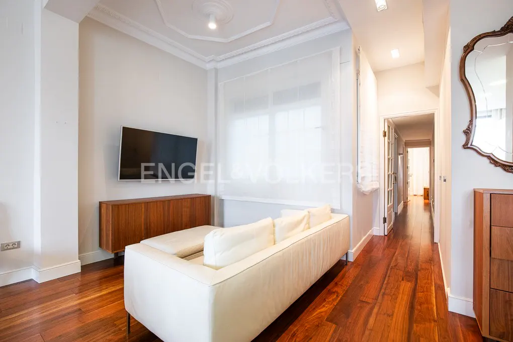 Bright living room with white sofa, wall-mounted TV, wooden sideboard, and polished hardwood floors leading to a hallway.