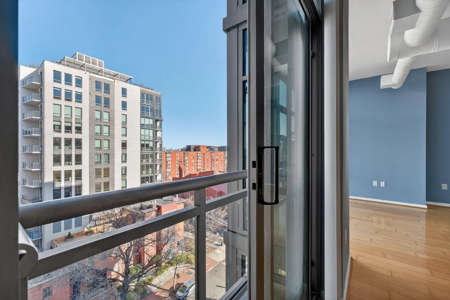 View from a condo balcony with a metal railing, overlooking city buildings under a blue sky. Inside, a blue wall and wood floor are visible.