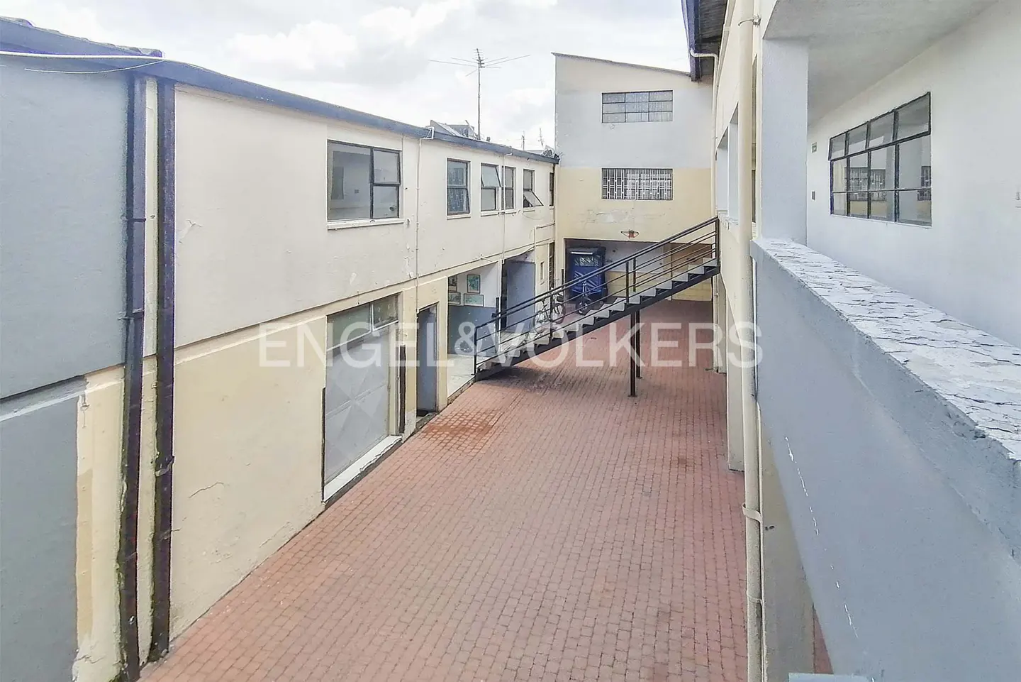 Exterior view of a commercial building with a brick courtyard and metal stairs. Windows and doors are visible on the building's facade.