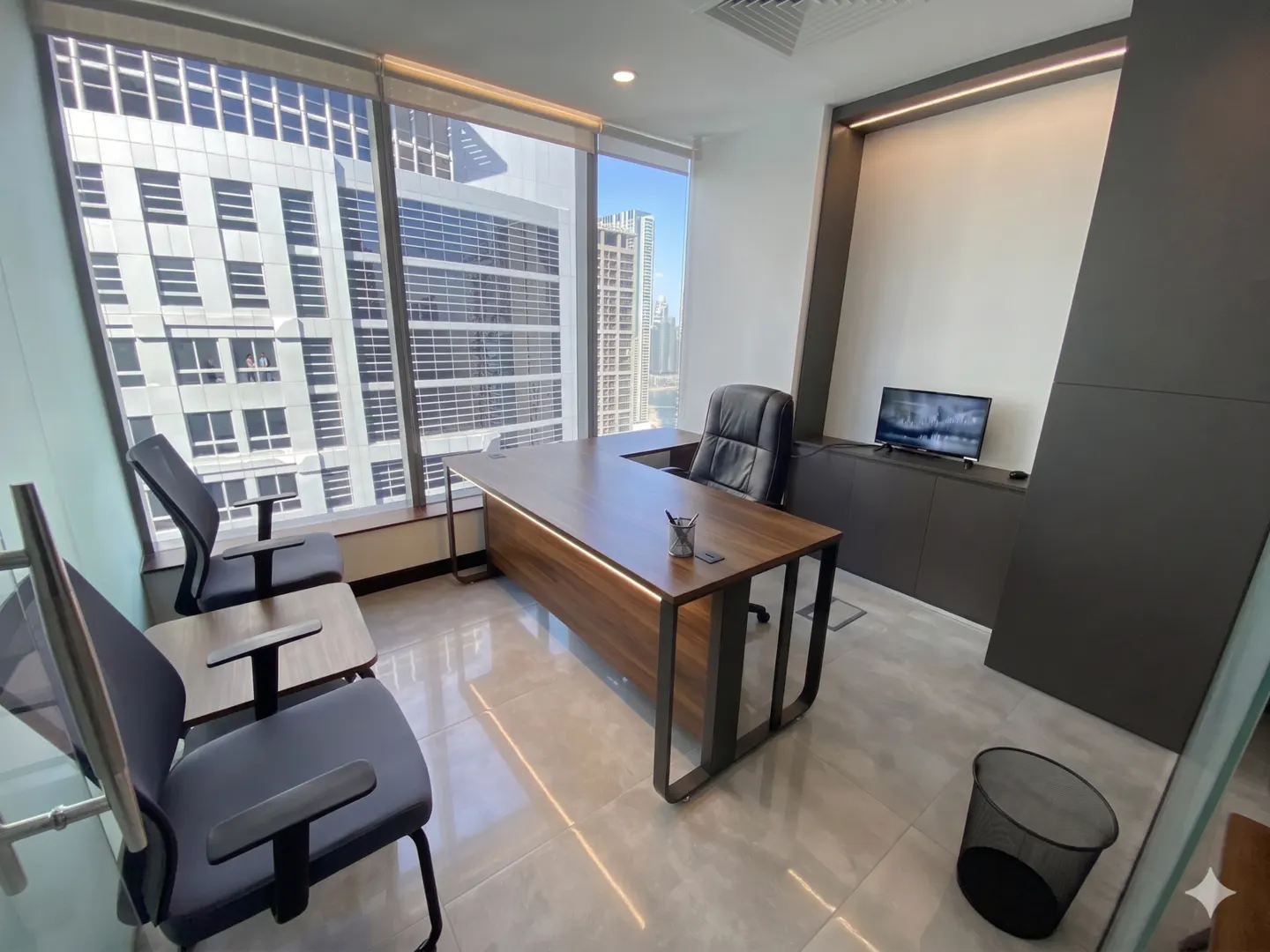 Modern office space with a large window view. A wooden desk, black leather chair, and guest seating are visible.
