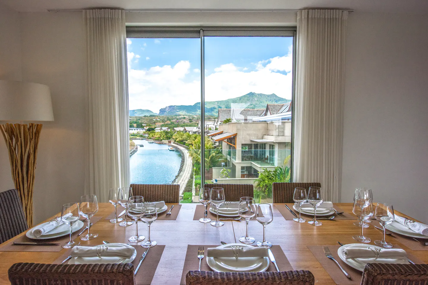Dining room with a set table, plates, glasses, and silverware. A large window shows a view of a canal, buildings, and mountains.
