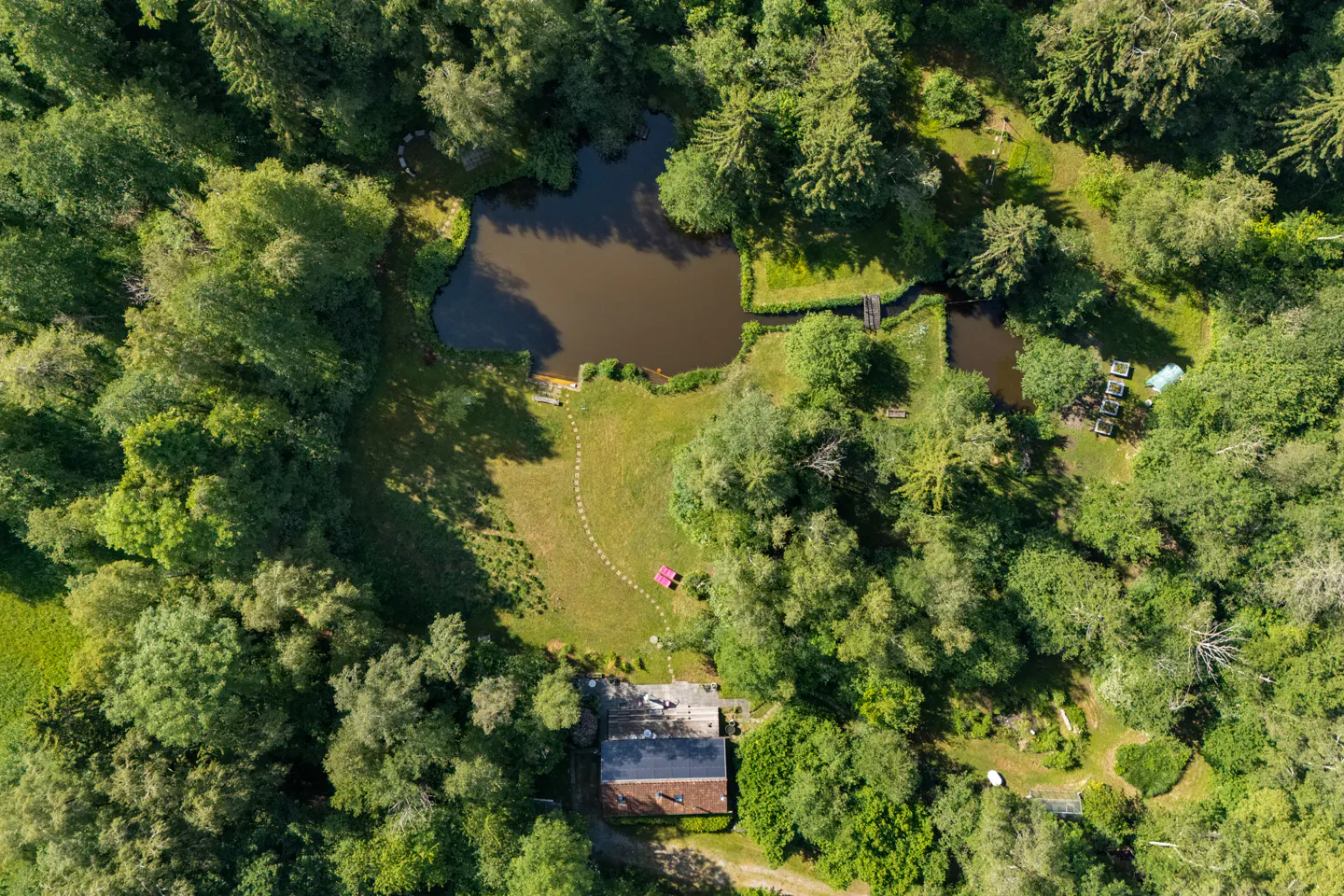 Aerial view of a house with a stone path leading to a pond, surrounded by dense green forest and lush landscaping.