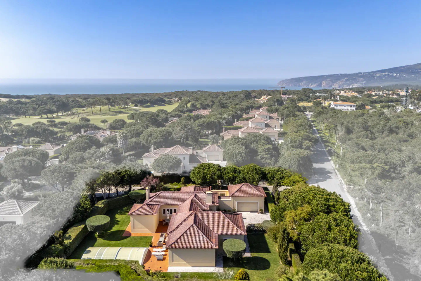 Aerial view of a tan house with a red tile roof, green lawn, and pool. Ocean and trees in the background.