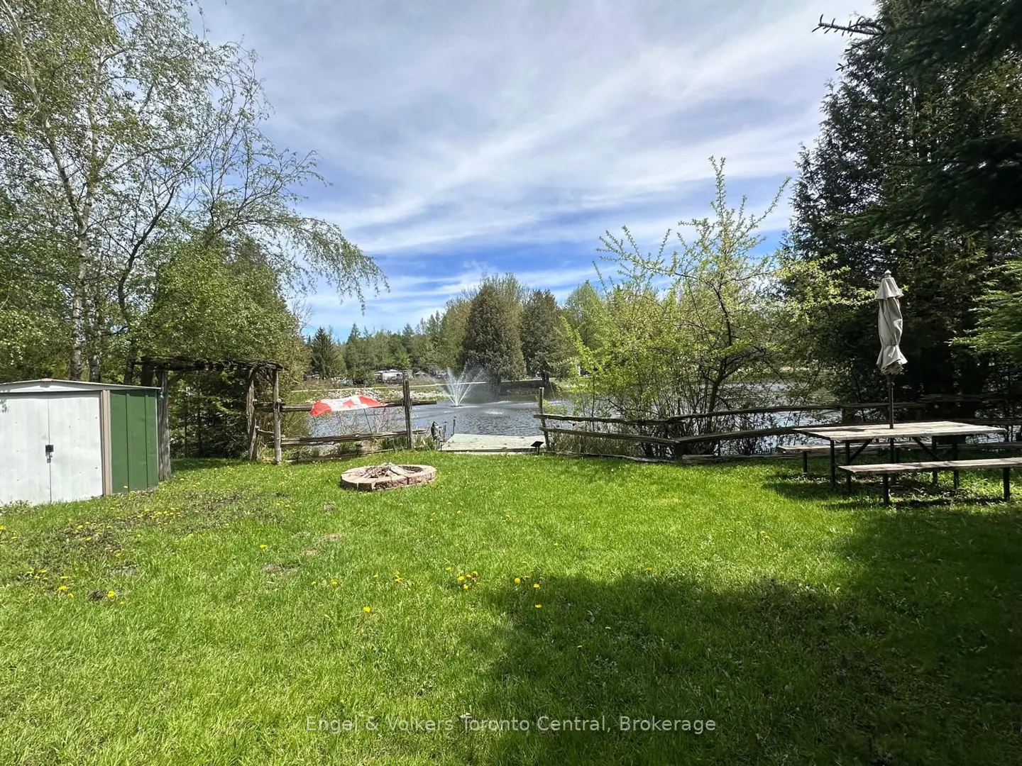 Lush green lawn with a fire pit, shed, and picnic table. A lake with a fountain is visible beyond a wooden fence. Trees and blue sky in the background.