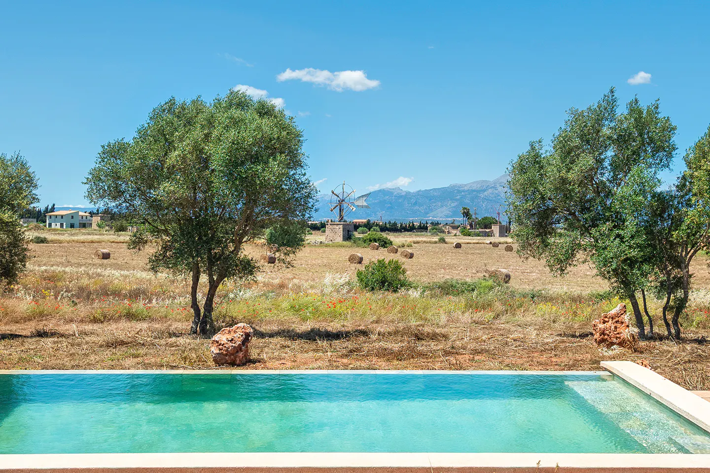 A turquoise pool in front of a field with hay bales, trees, and a windmill under a blue sky.