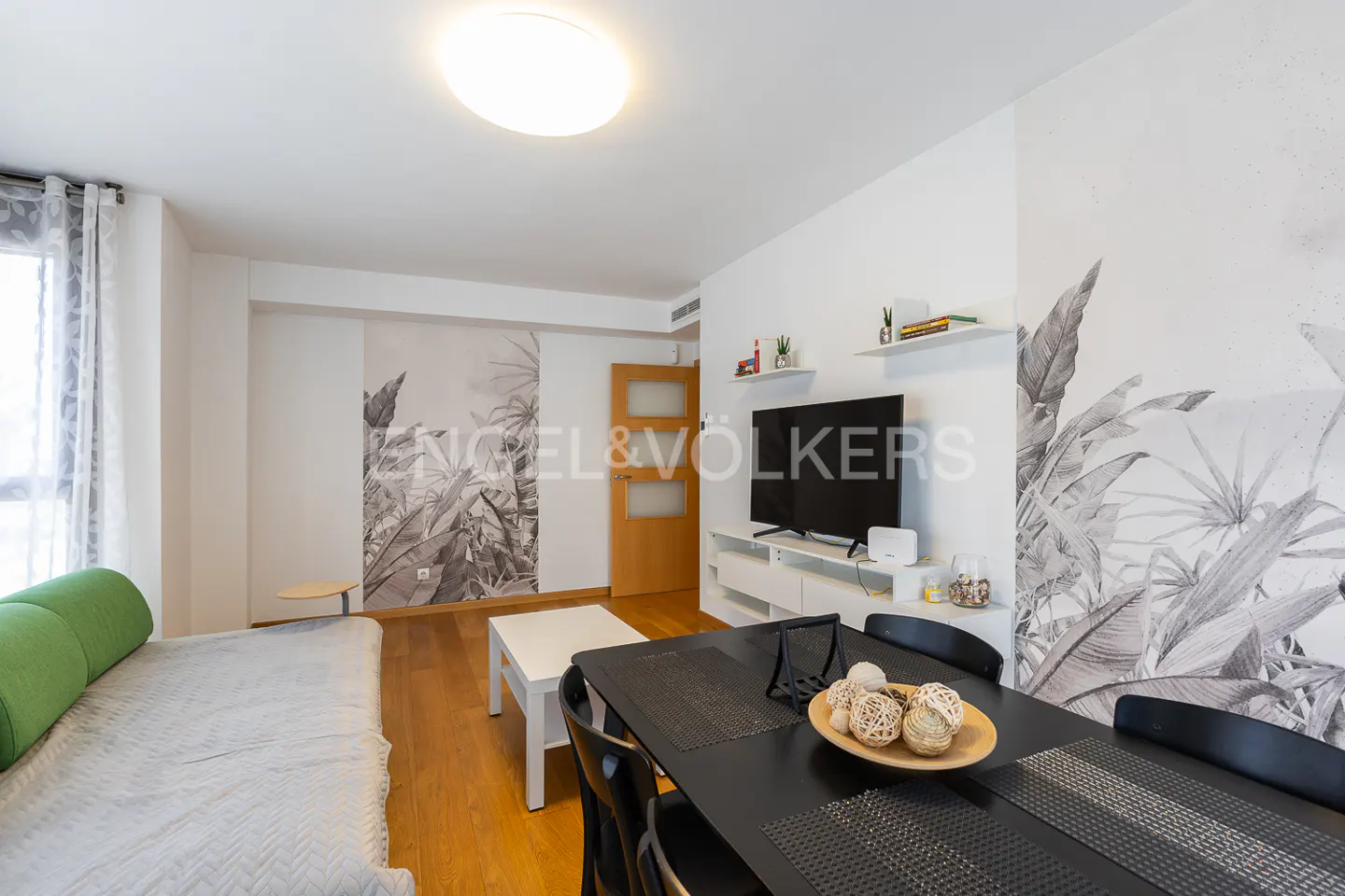 Bright living room with a black dining table, a white sofa with green pillows, and a TV. The walls have a tropical leaf pattern.