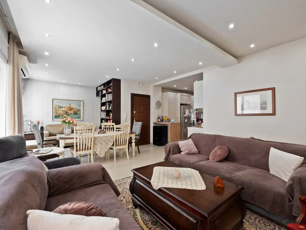 Bright living room with brown sofas, a dark wood coffee table, and a dining area with white chairs. A bookcase and kitchen are visible in the background.