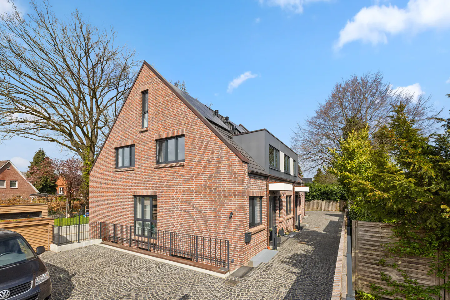 Exterior view of a two-story red brick house with black window frames and a cobblestone driveway under a blue sky.