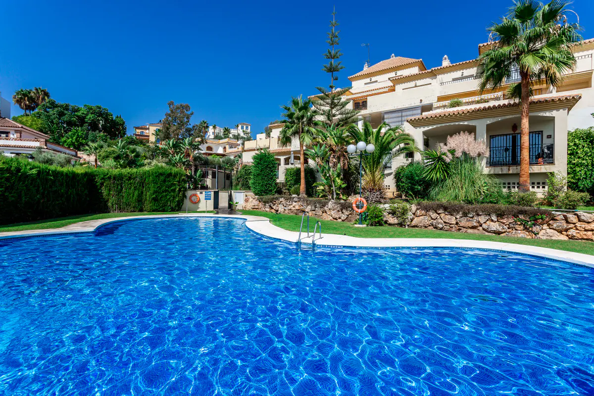 A bright blue swimming pool sits in front of a multi-story beige building with palm trees under a clear blue sky.