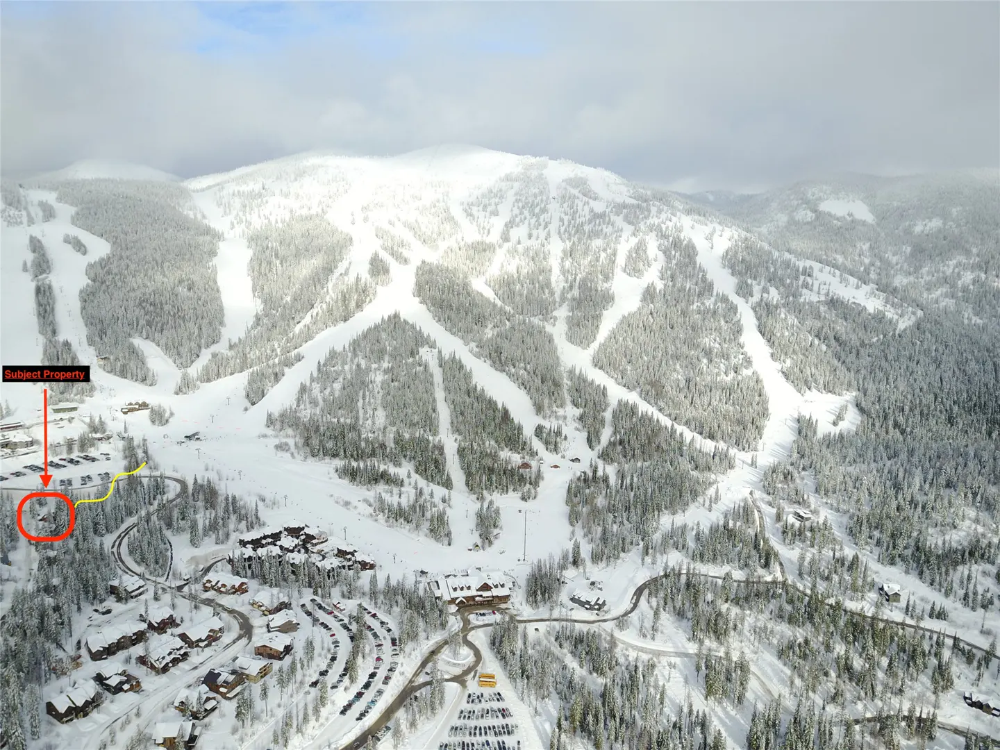 Aerial view of a snow-covered mountain with ski runs, trees, buildings, and parking lots. The subject property is circled in red.