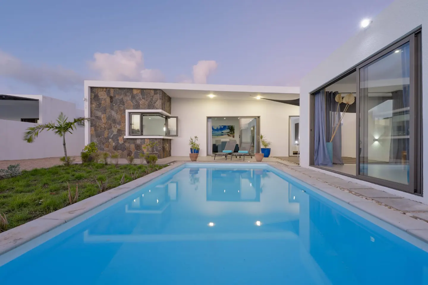 A modern home with a blue swimming pool in the foreground. The house has white walls and large windows. There are lounge chairs visible through the open sliding doors.