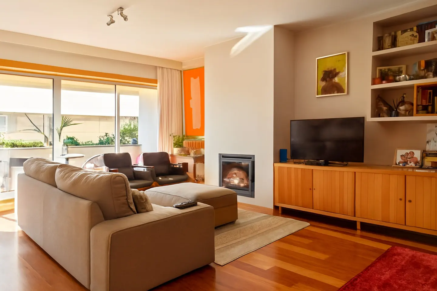 Living room with a beige sofa, two chairs, a fireplace, and a TV on a wooden cabinet. There is a balcony with plants in the background.