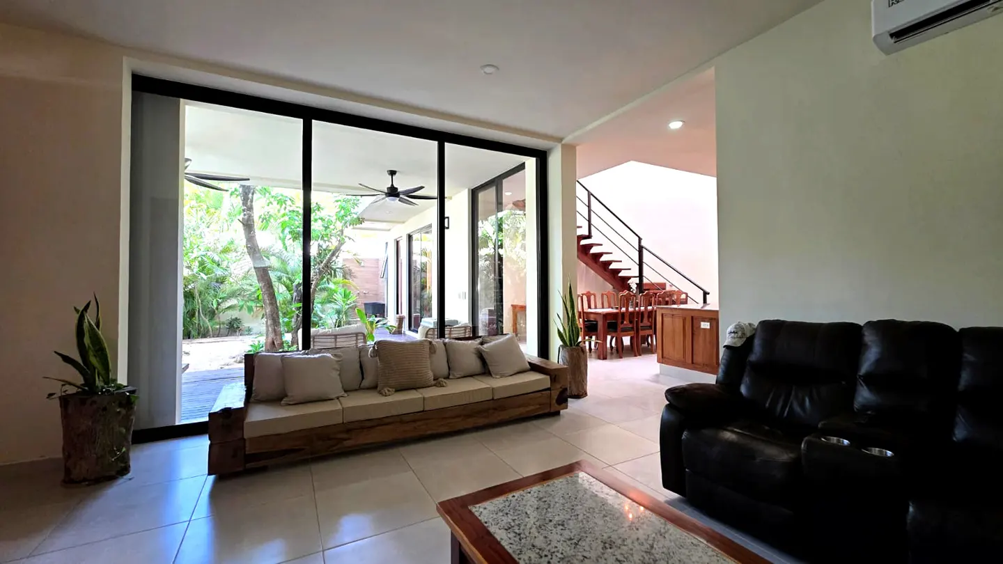 Bright living room with a wood sofa, black leather couch, and a view of a lush green outdoor space through large glass doors.