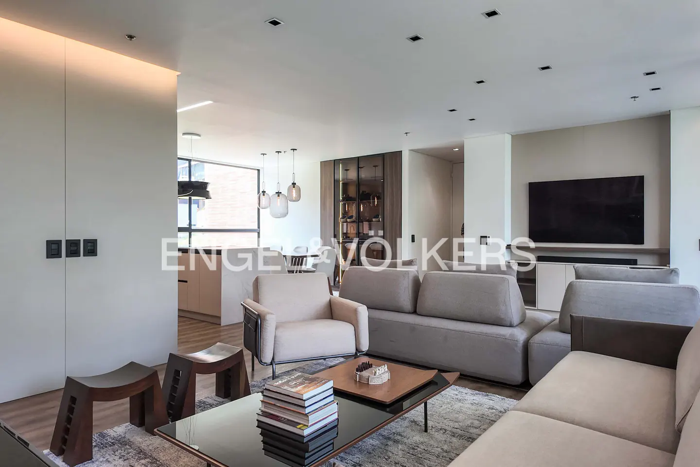 A modern living room with gray sofas, a glass coffee table with books, and wooden stools on a patterned rug. A TV is mounted on the wall.